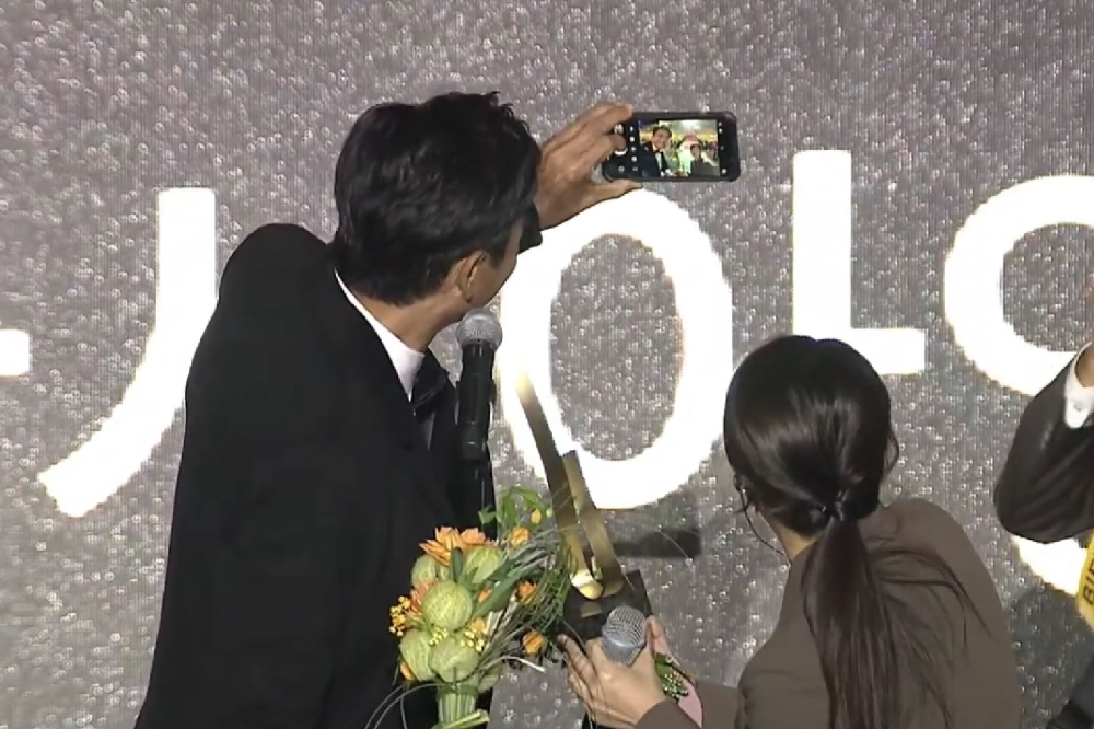 周润发说完感言后，突然将奖座递给翻译员，随即从西装拿出手机，要跟台下观众大合照。-第28届釜山国际电影节开幕礼视频截图-
