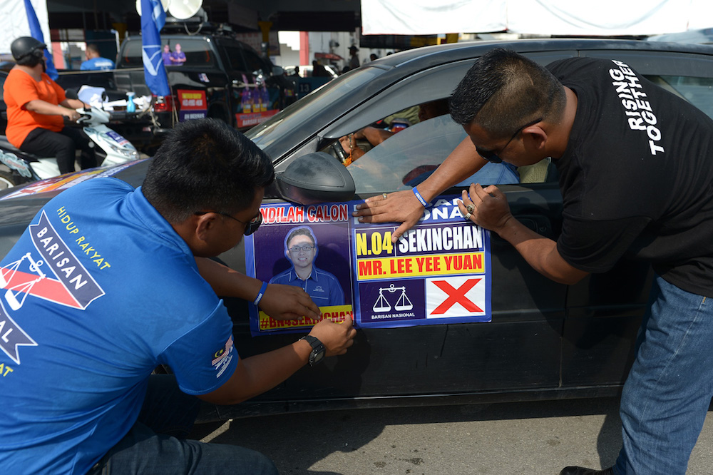 Two men, including one wearing a BN shirt, are seen pasting a sticker featuring BN's Sekinchan candidate on a car. u00e2u20acu201d Picture by Mukhriz Hazim