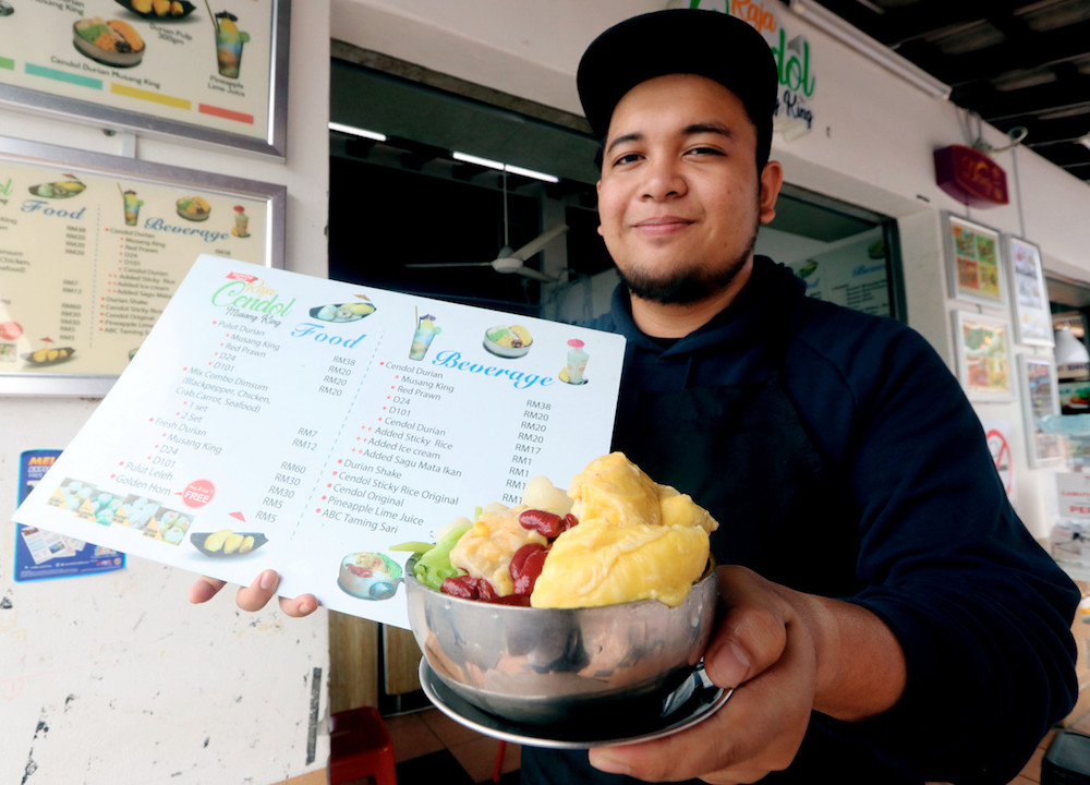 Raja Cendol Taming Sari owner Muhammad Ariff Qayyum Mohd Zamri poses for pictures with a bowl of Musang King cendol and a menu at his stall in Bandar Hilir, Melaka July 12, 2019. u00e2u20acu201d Bernama pic
